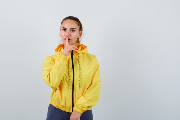Young lady in yellow jacket showing silence gesture and looking confident , front view.