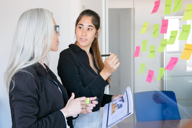 Young Latin employer holding pen and statistic documents and looking at boss. Focused confident grey-haired female manager training colleague