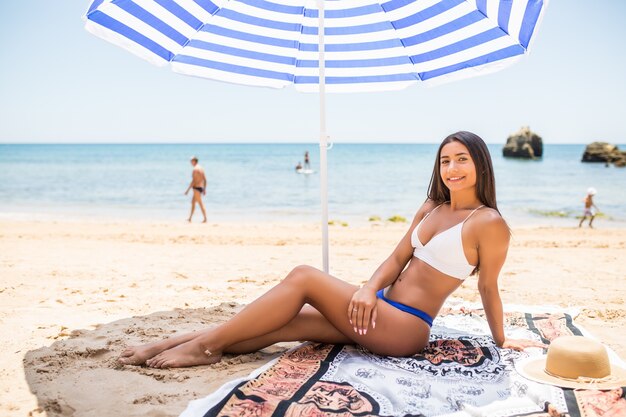 Young latin girl in bikini lying under color umbrella on the beach in sunny hot day.