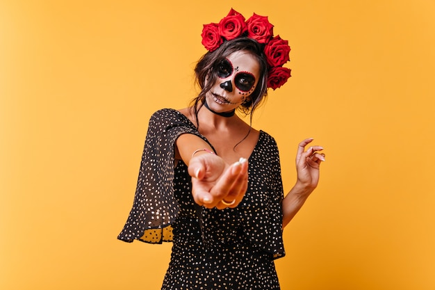 Young latin model calls by hand to join her. portrait of dark-haired woman in image of skeleton on isolated wall