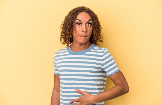 Young latin transsexual woman isolated on yellow background touches tummy, smiles gently, eating and satisfaction concept.