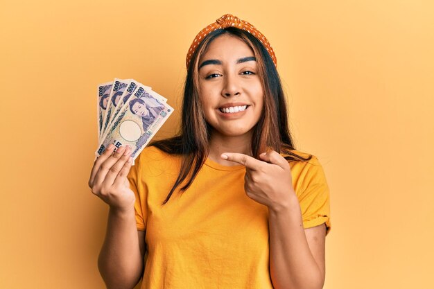 Young latin woman holding japanese yen banknotes smiling happy pointing with hand and finger