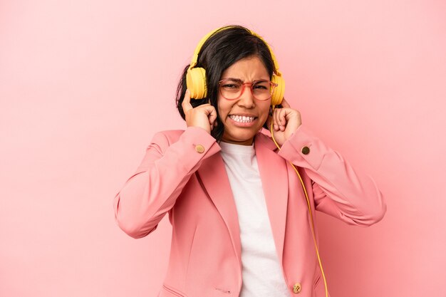 手で耳を覆うピンクの背景に分離された音楽を聴いている若いラテン女性。