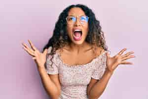 Foto gratuita giovane donna latina che indossa abiti casual e occhiali pazzi e pazzi che gridano e urlano con espressione aggressiva e braccia alzate. concetto di frustrazione.