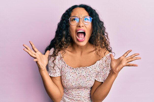 Young latin woman wearing casual clothes and glasses crazy and mad shouting and yelling with aggressive expression and arms raised. frustration concept.