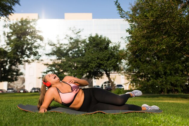 Молодая смеющаяся женщина больших размеров в спортивном топе и леггинсах с красными наушниками лежит на коврике для йоги, счастливо проводя время в городском парке
