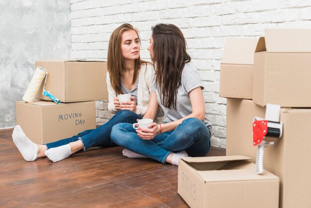 Young lesbian couple holding coffee cup in hands looking at each other sitting among the cardboard boxes