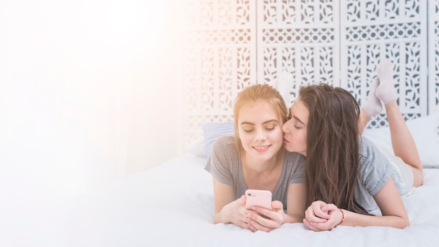 Young lesbian woman lying on bed kissing her girlfriend using mobile phone