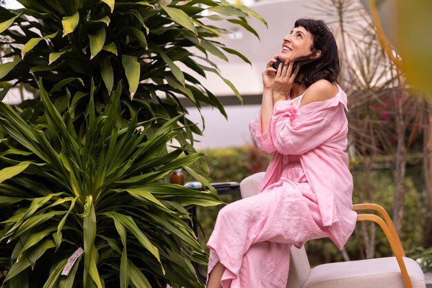 Young lightskinned brunette girl in casual clothes looks up using phone sitting in tropical garden during day Lifestyle concept