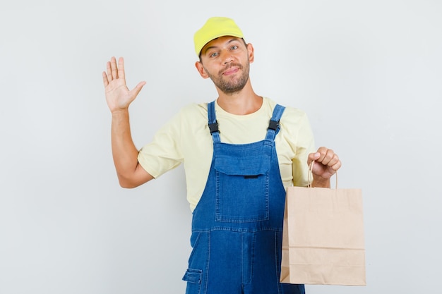 Young loader holding paper bag and waving hand in uniform and looking cheerful. front view.