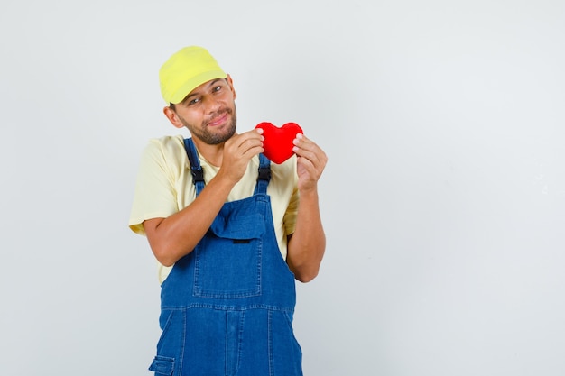 Young loader holding red heart in uniform and looking joyful , front view.