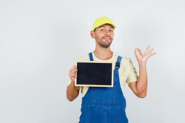 Young loader in uniform holding blackboard with ok sign and looking cheerful , front view.