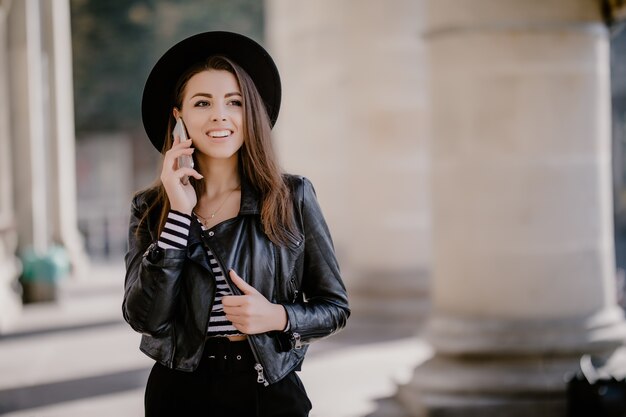 가죽 재킷에 젊은 사랑스러운 갈색 머리 소녀, 도시 산책로의 검은 모자는 전화 대화를