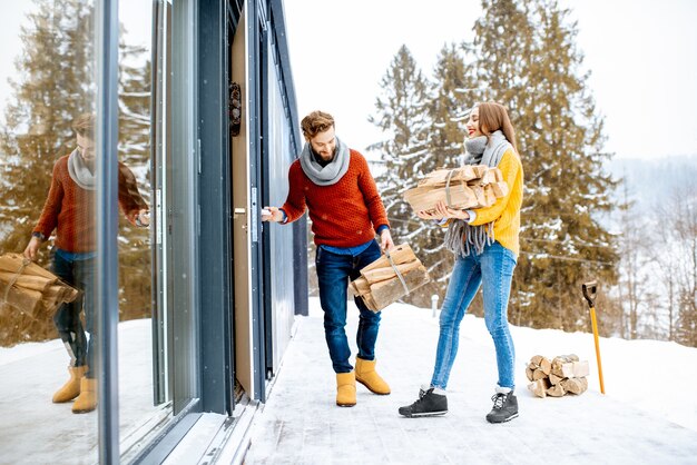 Молодая милая пара, одетая в красочные свитера, входит в свой современный дом с дровами в горах зимой