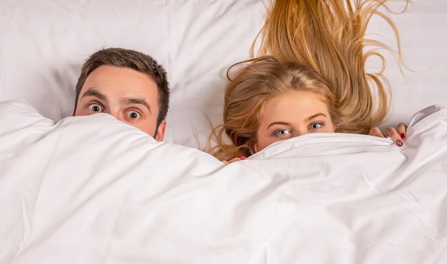 Молодая милая пара лежит и веселится в постели, счастливая улыбка смотрит в камеру, укрывается одеялом. Концепция семьи, времени сна и счастья