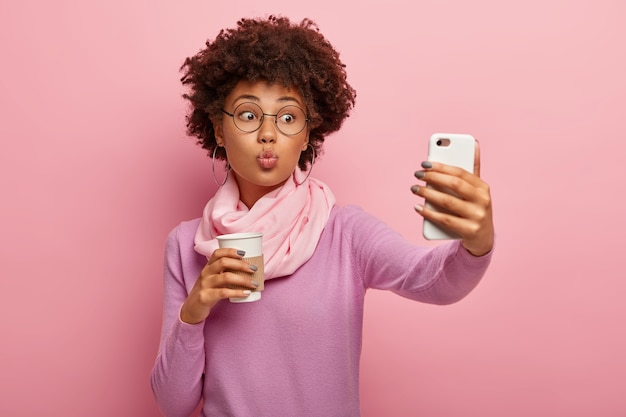 Young lovely woman with afro haircut, keeps lips folded, blows kiss at camera of smartphone, takes selfie portrait