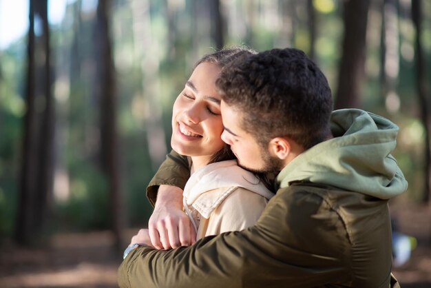 Young loving couple hugging in forest. Man hugging and kissing smiling woman. Love, affection, relationship concept