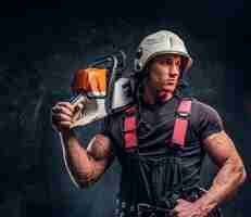 Free photo young lumberjack wearing protective clothes posing with a chainsaw and looks sideways in a dark studio