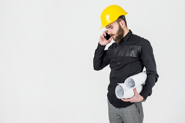 Young male architect holding blue prints talking on mobile phone