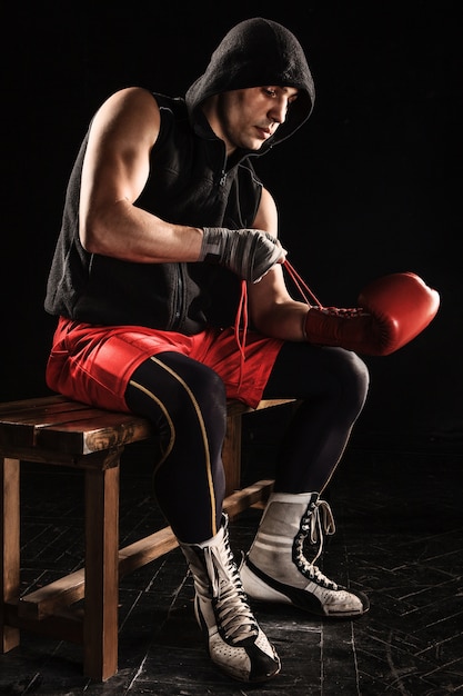 young male athlete kickboxing sitting and lacing glove on a black background