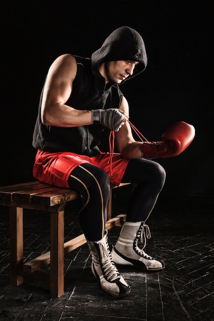 The young male athlete kickboxing sitting and lacing glove on a black 