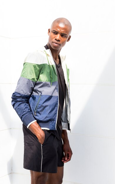 Young male athlete with hands in his pocket standing against white wall