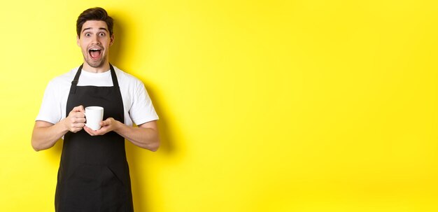 Young male barista holding coffee cup and looking surprised standing in black apron against yellow b