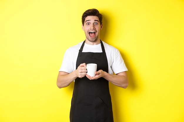 黄色の背景に黒いエプロンで立って、コーヒーカップを保持し、驚いて見える若い男性バリスタ