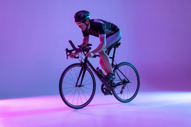 Young male bike rider on bicycle isolated on gradient wall in neon man training and practicing