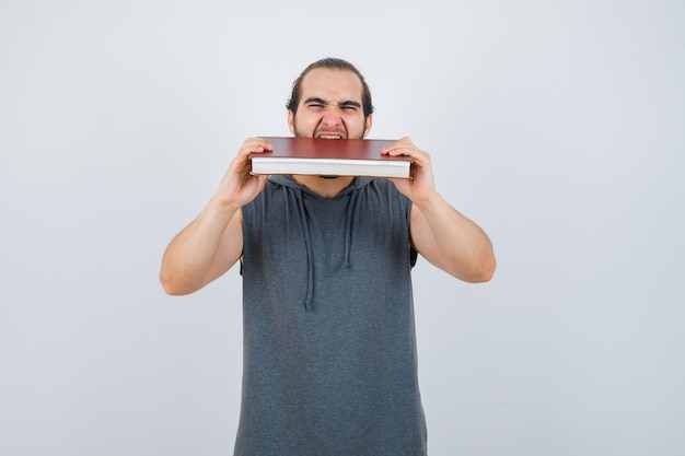 Free photo young male biting book in sleeveless hoodie and looking ravenous. front view.