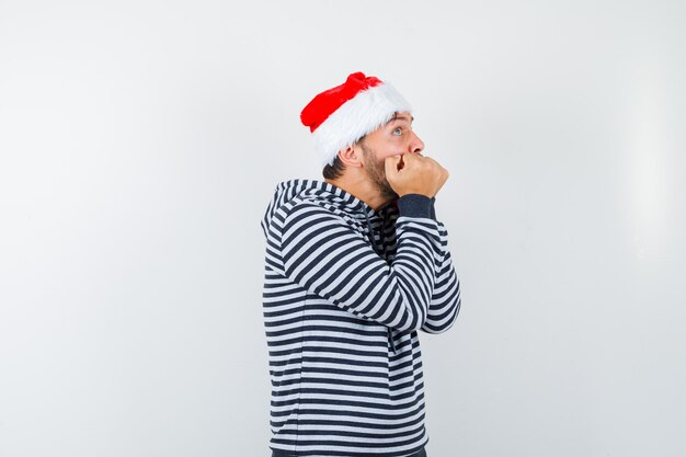 Young male biting fists emotionally in hoodie, Santa hat and looking frightened ,