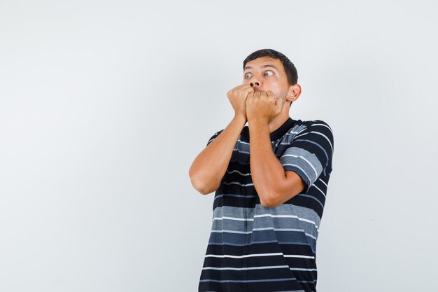 Young male biting fists in t-shirt and looking frightened , front view.