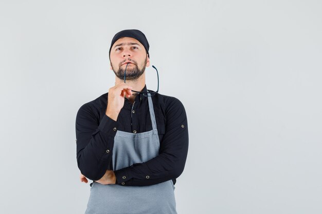 Young male biting glasses while looking up in shirt, apron and looking pensive. front view.