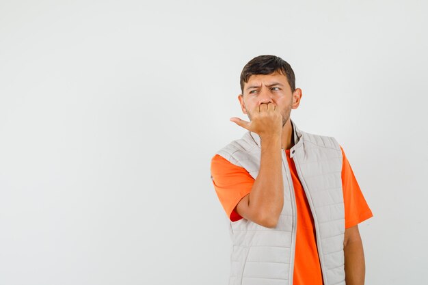 Young male biting nails while looking aside in t-shirt, jacket and looking troubled. front view.