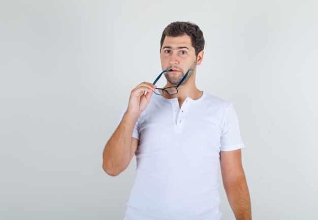 Young male biting slightly his glasses in white t-shirt and looking thoughtful