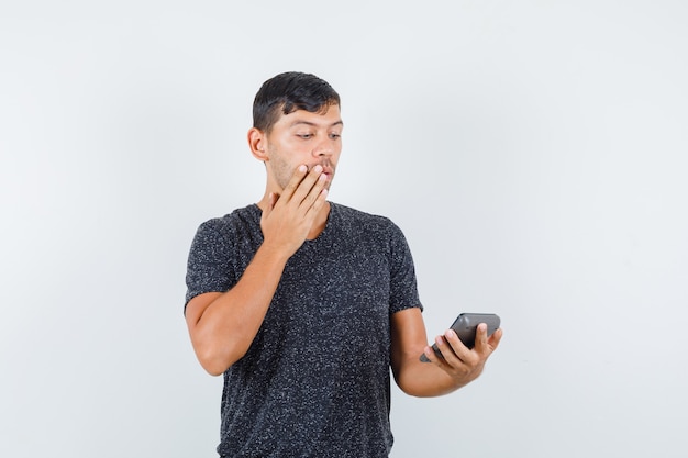 Young male in black t-shirt looking at calculator and looking surprised , front view.