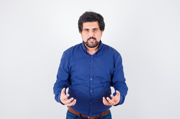 Young male in blue shirt keeping hands in aggressive manner and looking angry , front view.