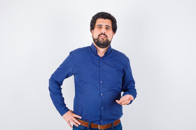 Young male in blue shirt stretching hand in puzzled gesture and looking confident , front view.