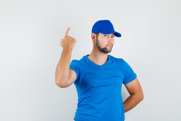Young male in blue t-shirt and cap pointing up with hand on back and looking sly