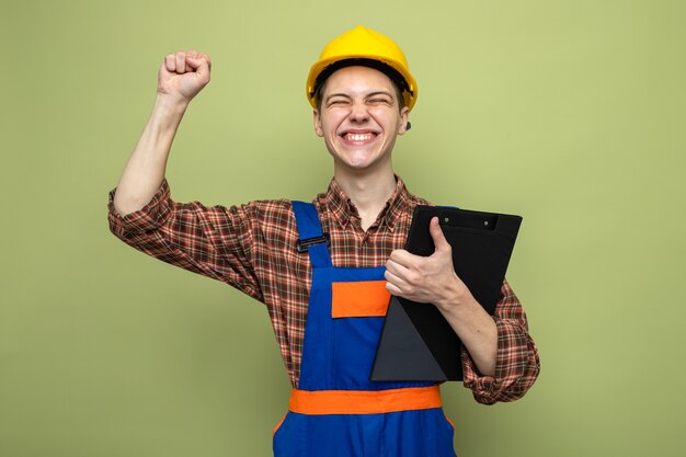 Free photo young male builder holding clipboard wearing uniform isolated on olive green wall