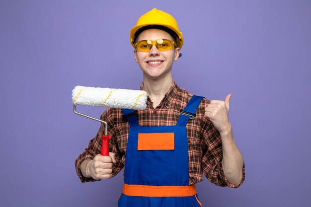 コピースペースと紫色の壁に分離されたローラーブラシを保持している制服と眼鏡を身に着けている若い男性ビルダー