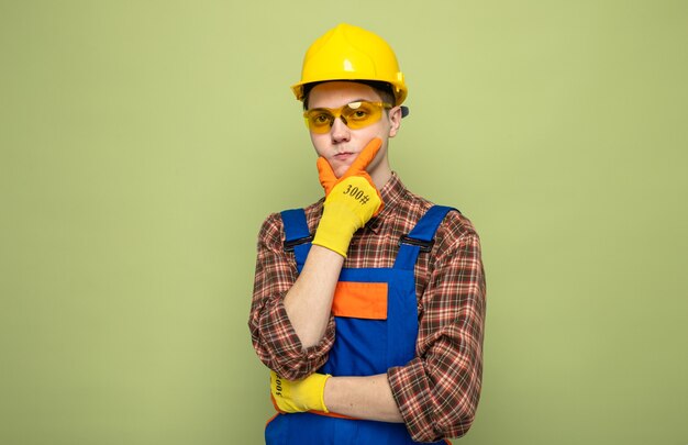 Young male builder wearing uniform and gloves with glasses isolated on olive green wall