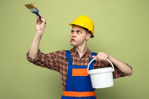 Free photo young male builder wearing uniform holding bucket with paint brush isolated on olive green wall