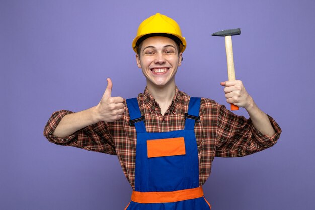 Young male builder wearing uniform holding hammer isolated on purple wall