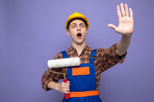 Молодой мужчина-строитель в униформе держит роликовую щетку, изолированную на фиолетовой стене