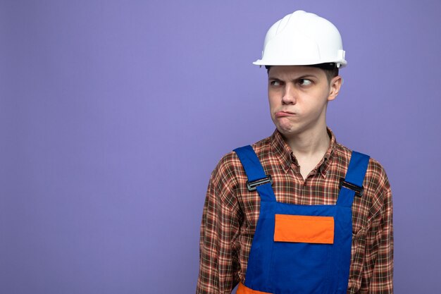 Young male builder wearing uniform isolated on purple wall with copy space