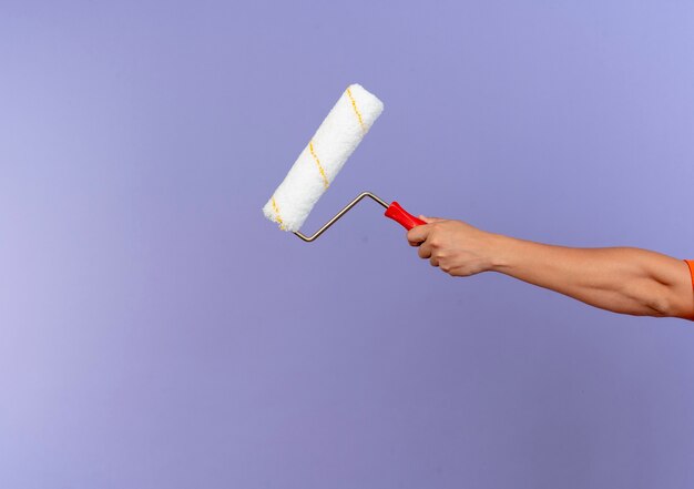 Young male builder wearing uniform and safety helmet holding paint roller isolated on purple wall