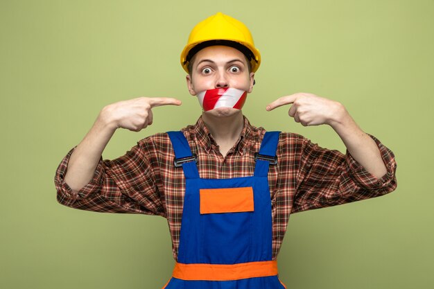 Young male builder wearing uniform sealed mouth with duct tape isolated on olive green wall