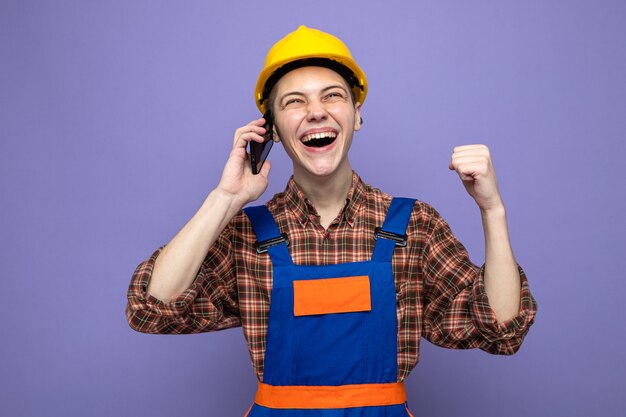 Young male builder wearing uniform speaks on phone isolated on purple wall