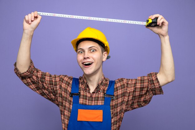 Young male builder wearing uniform stretching out measure tape isolated on purple wall